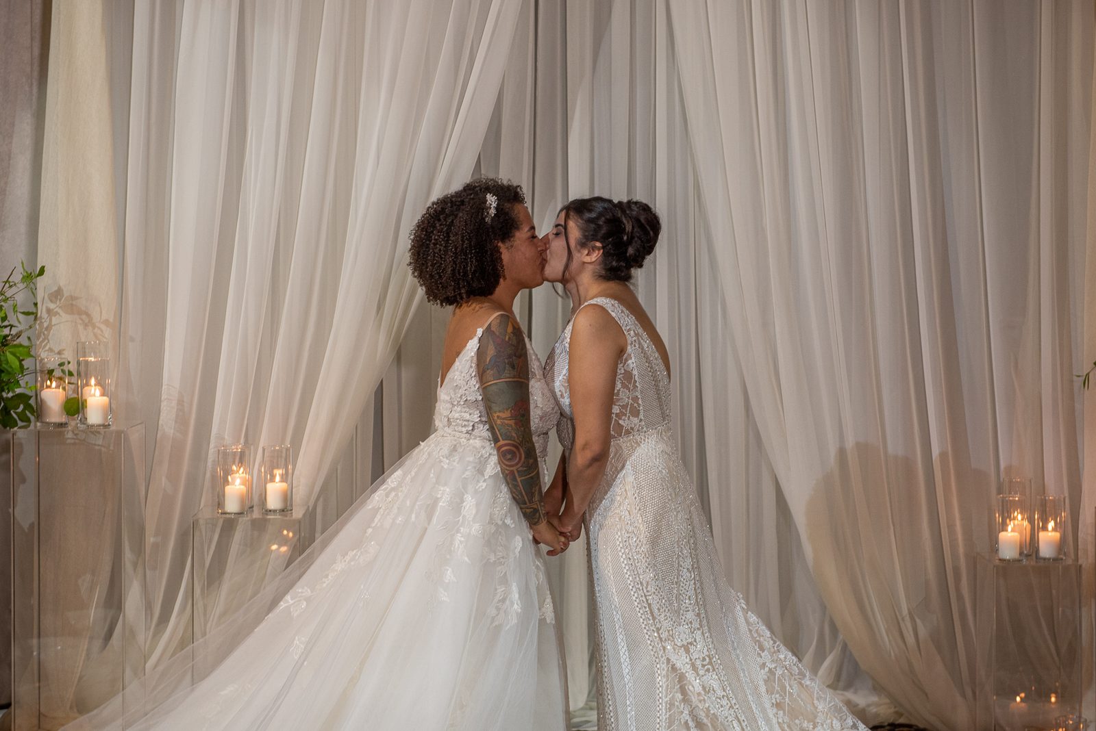 Two brides with flowing drapery behind them and candles on clear pedestals share a tender kiss while holding hands.