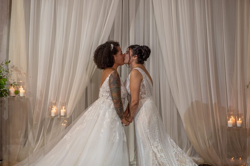 Two brides with flowing drapery behind them and candles on clear pedestals share a tender kiss while holding hands.