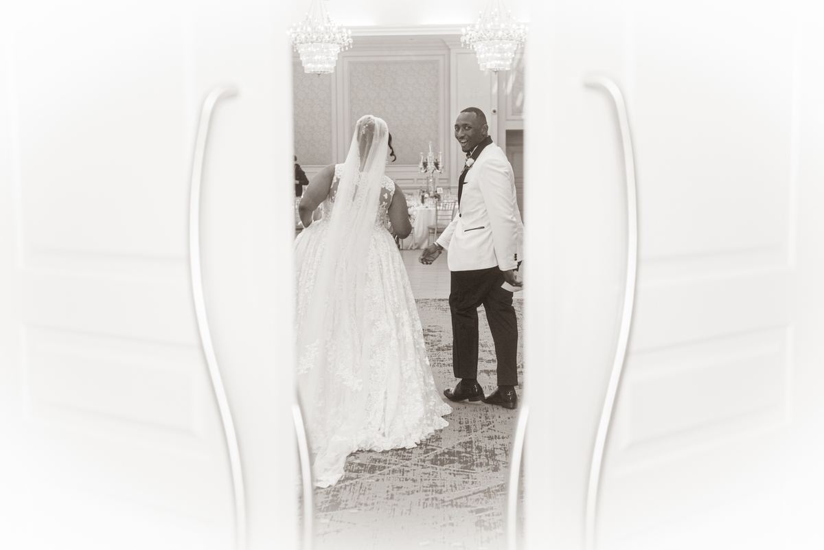 Groom in white tuxedo smiles widely as bride in lace gown and veil walks toward him, illuminated by soft chandelier light.