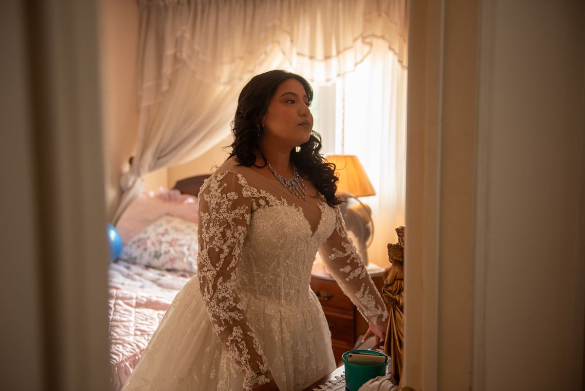 A bride in a lace gown stands by a window, gazing thoughtfully as sunlight filters through sheer curtains.