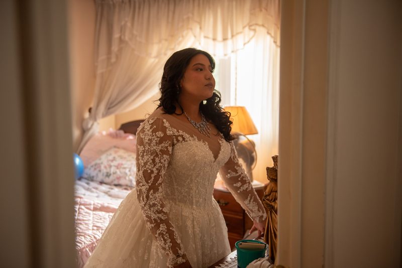 A bride in a lace gown stands by a window, gazing thoughtfully as sunlight filters through sheer curtains.