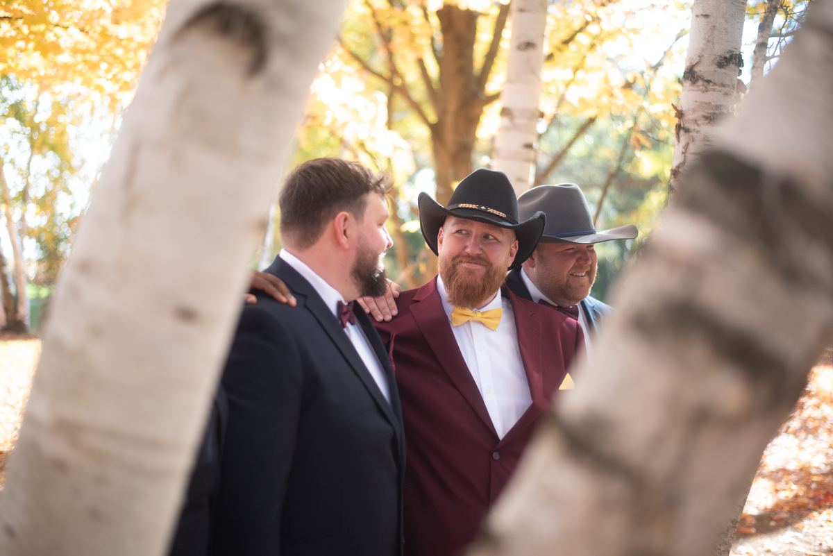 Three formally dressed men in cowboy hats stand joyfully together in an autumn grove, illuminated by warm sunlight.