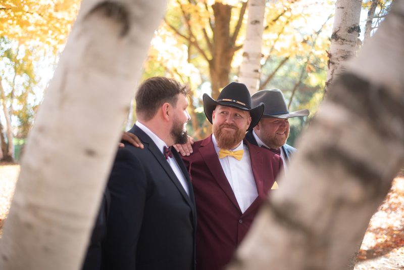 Three formally dressed men in cowboy hats stand joyfully together in an autumn grove, illuminated by warm sunlight.