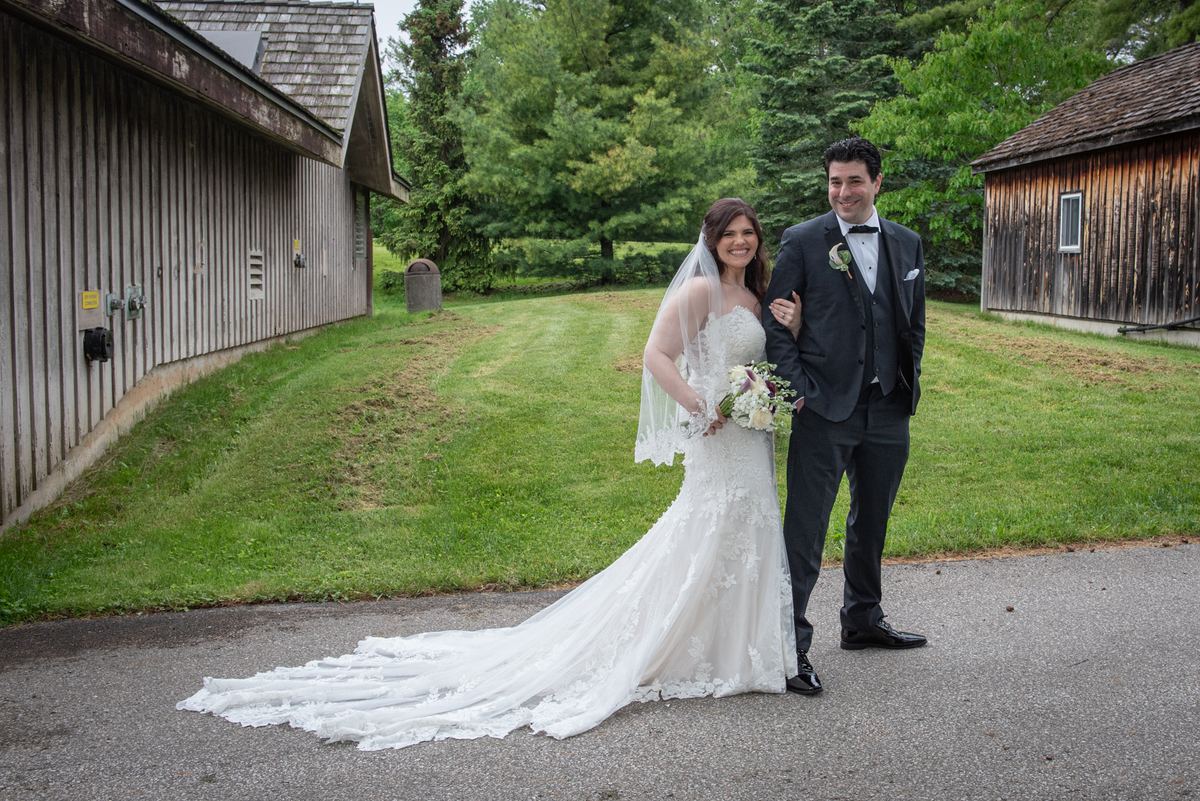 A smiling bride in a lace-trimmed gown and veil stands with her arm around the groom's waist, both surrounded by greenery.