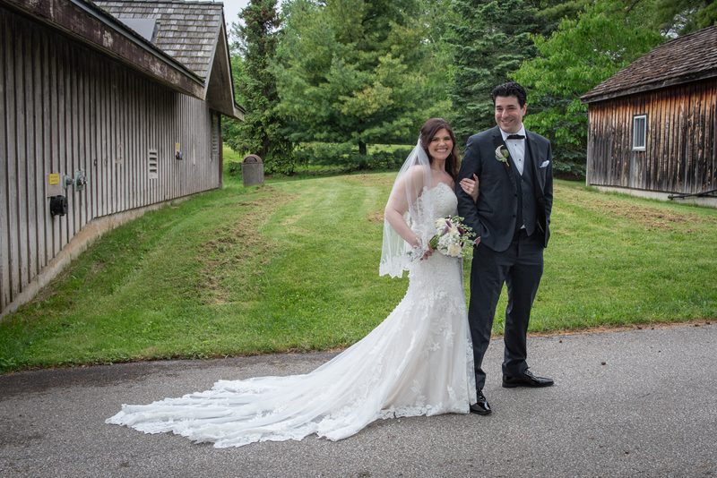 A smiling bride in a lace-trimmed gown and veil stands with her arm around the groom's waist, both surrounded by greenery.