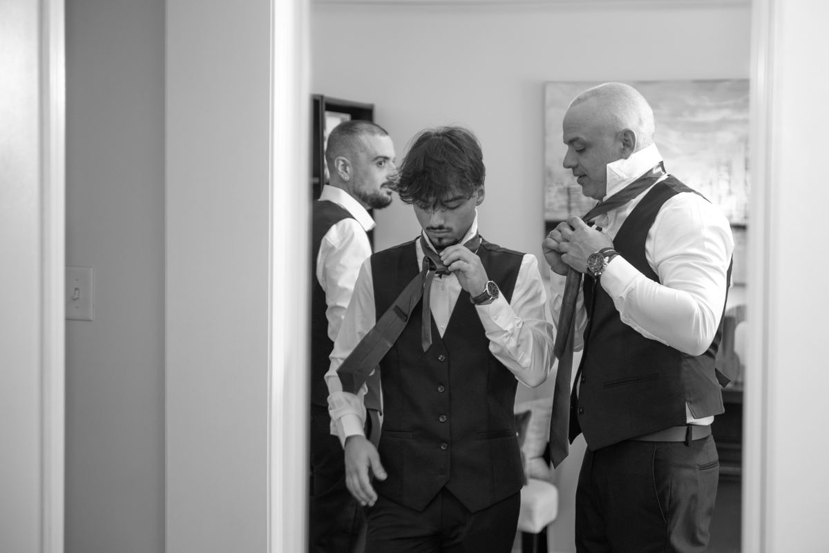 In an intimate indoor setting, a groom is assisted by an older man adjusting his bow tie while another man watches.