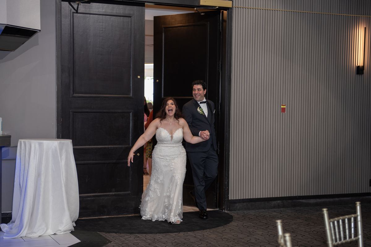 A bride and groom laugh joyfully as they enter a banquet hall through dark wood doors, holding hands.