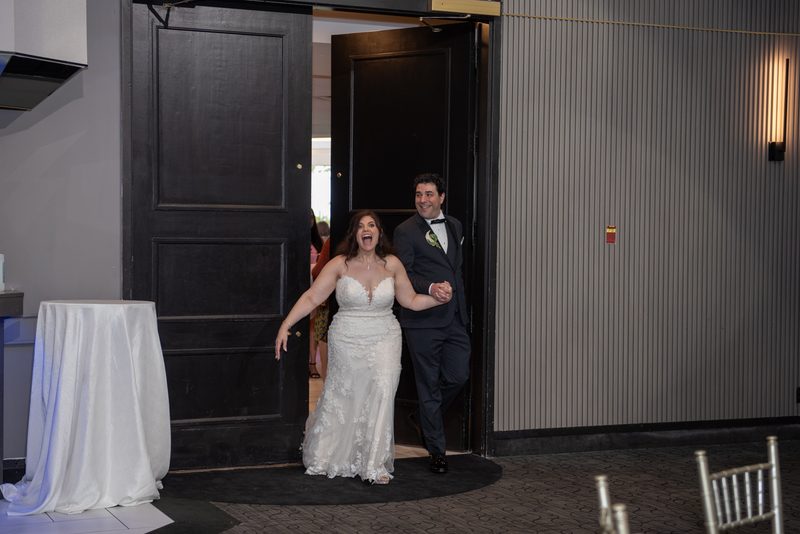 A bride and groom laugh joyfully as they enter a banquet hall through dark wood doors, holding hands.