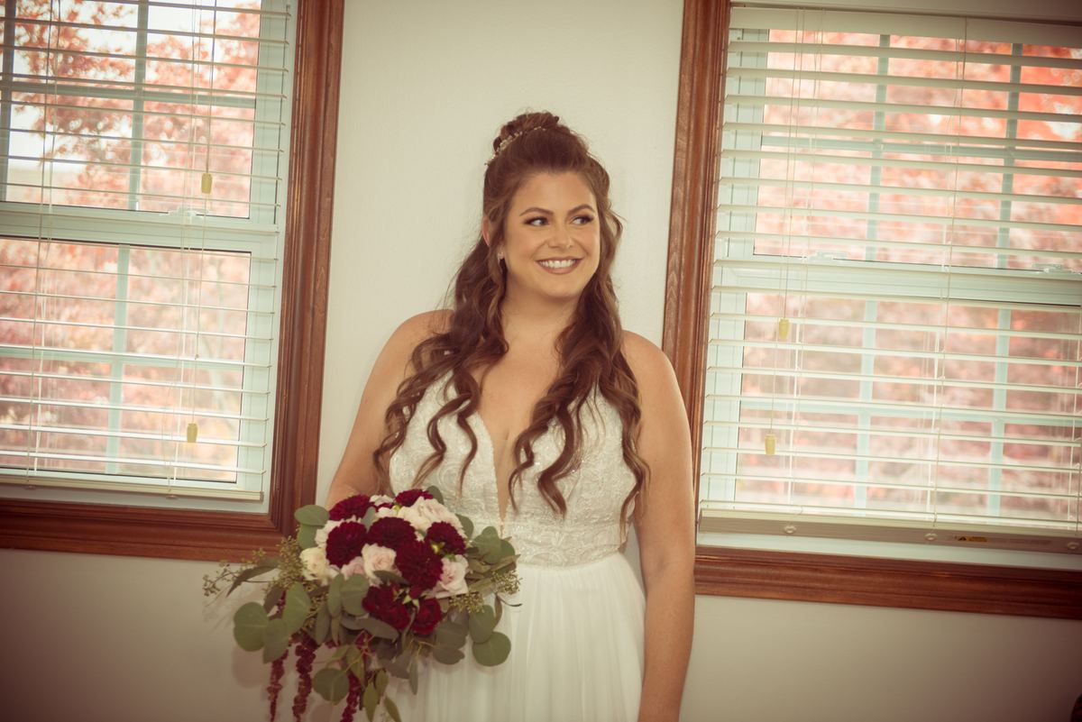 A smiling bride in a white lace gown holds a bouquet of red and pink flowers near two windows with blinds.