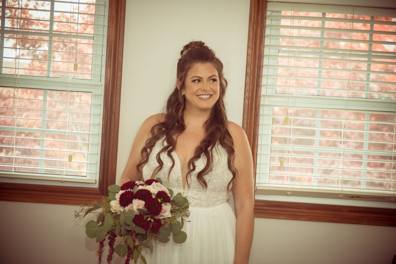 A smiling bride in a white lace gown holds a bouquet of red and pink flowers near two windows with blinds.
