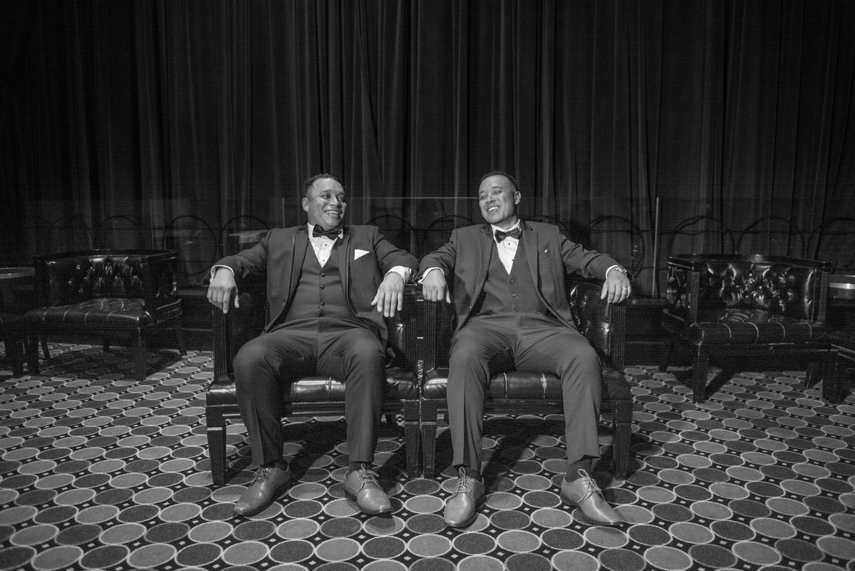 Two men in tuxedos laugh together in an elegant, dimly lit room with patterned carpet and heavy curtains.