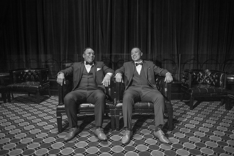 Two men in tuxedos laugh together in an elegant, dimly lit room with patterned carpet and heavy curtains.