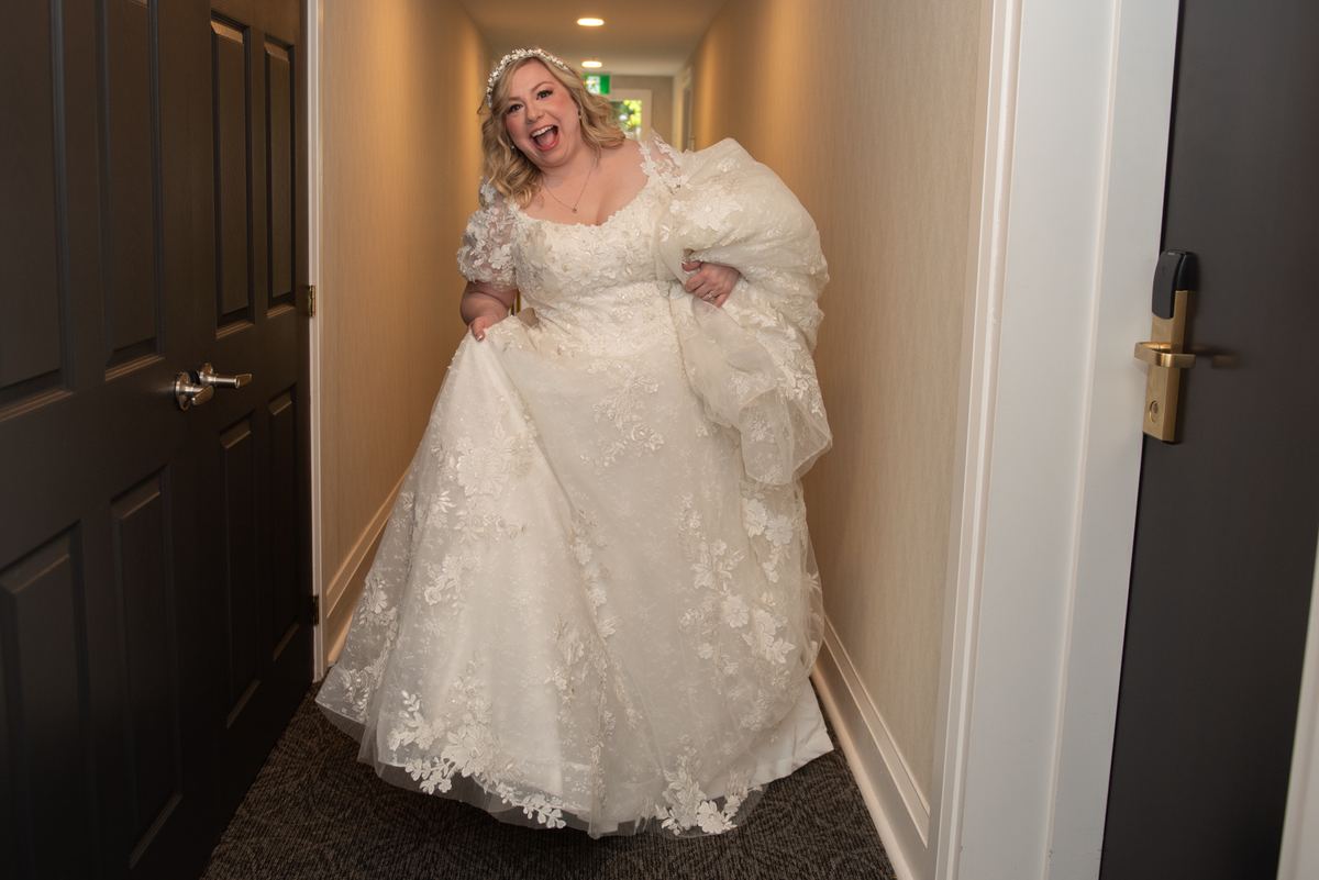 A joyful bride in a voluminous lace wedding gown laughs exuberantly as she bursts through a doorway into a hallway.