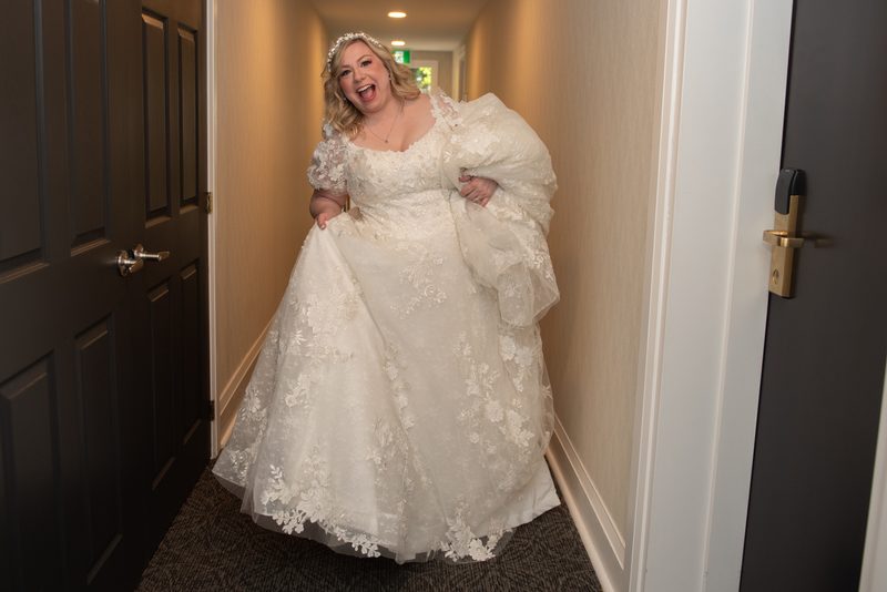 A joyful bride in a voluminous lace wedding gown laughs exuberantly as she bursts through a doorway into a hallway.