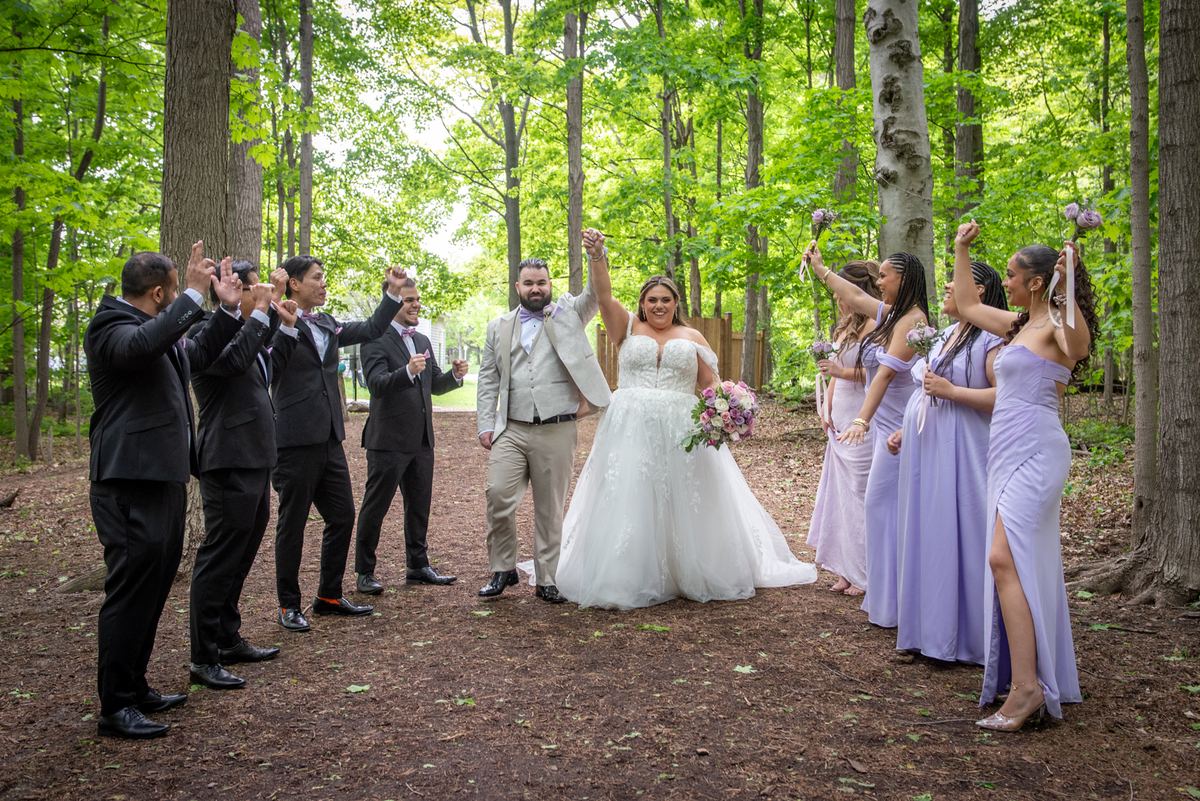 Bride and groom raise hands in celebration with joyful wedding party surrounding them in forest clearing.