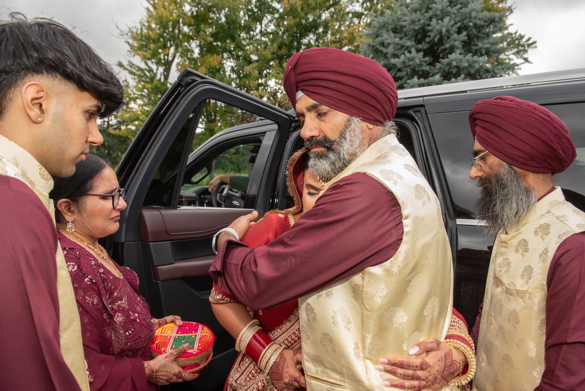 A Sikh groom in maroon turban embraces his bride stepping from a car, surrounded by family members in traditional attire.
