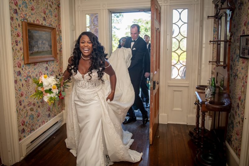 A smiling bride in a white lace gown walks through an ornate doorway into a warmly lit room, trailing her train.