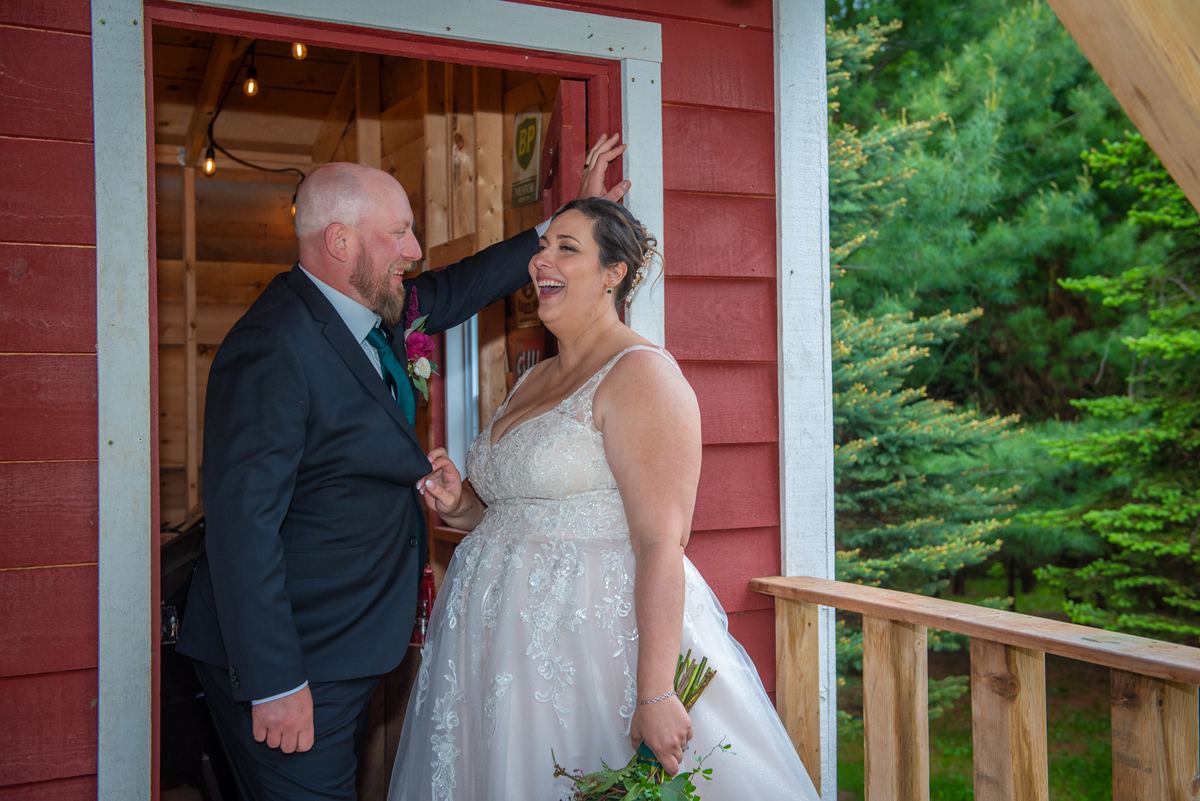 A laughing bride and groom stand in a doorway surrounded by trees, the groom playfully touching her head.