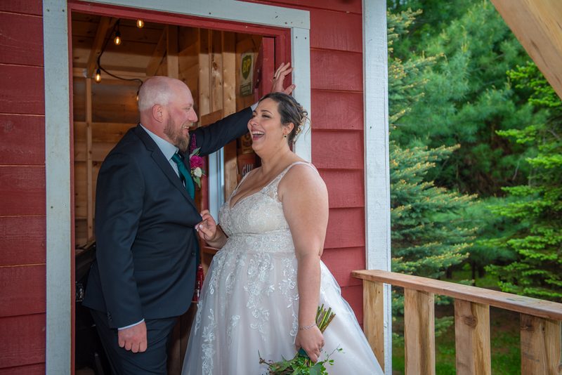 A laughing bride and groom stand in a doorway surrounded by trees, the groom playfully touching her head.