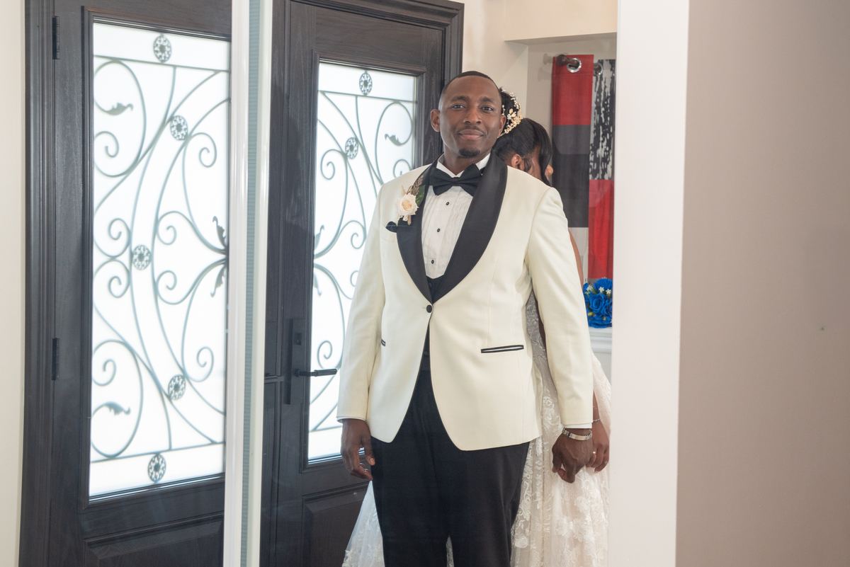 A groom in a white tuxedo stands in a doorway, looking expectantly at the camera as his bride holds his hand beside him.