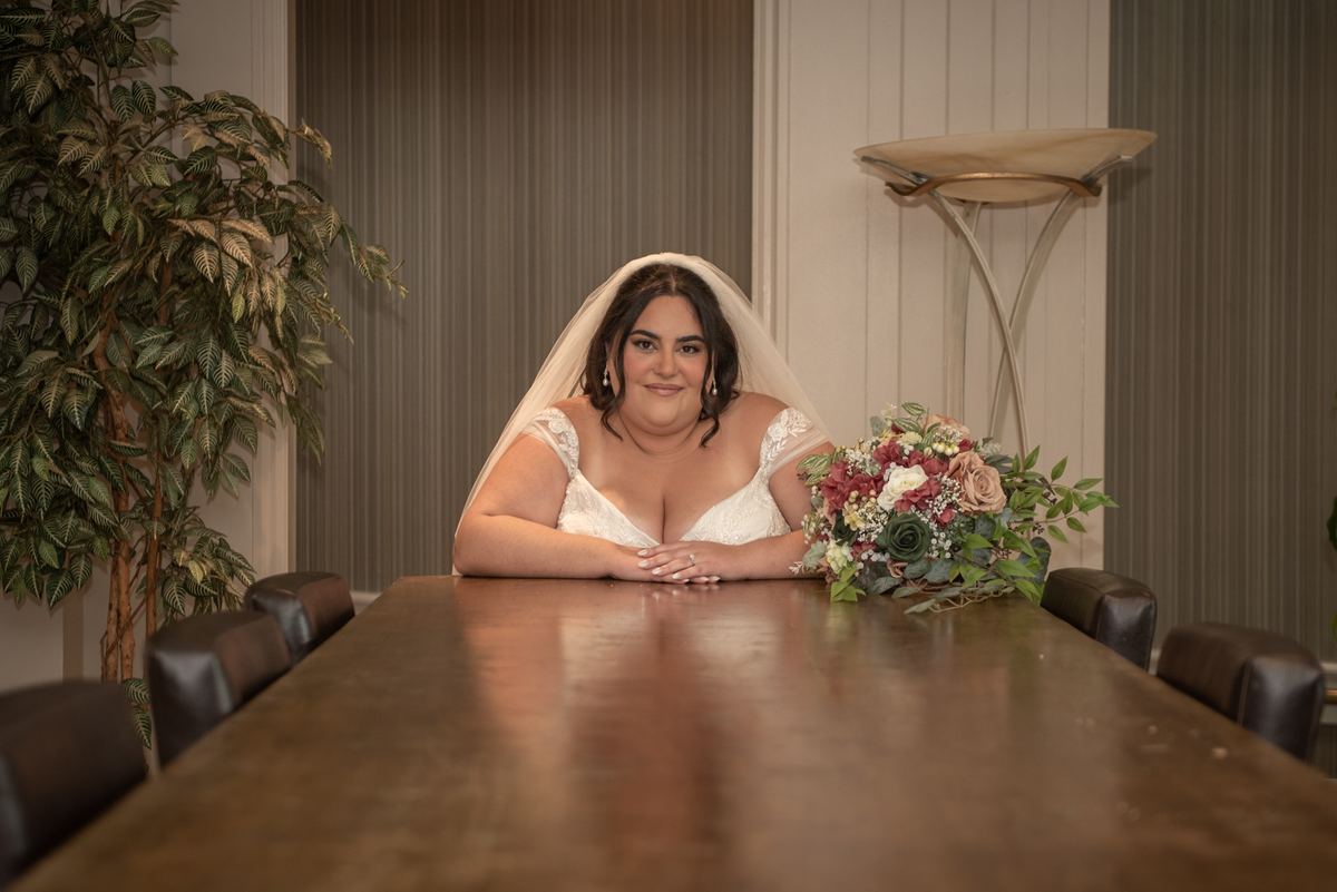 A bride in a white lace gown and veil smiles beside a wooden table with flowers, under ambient indoor lighting.