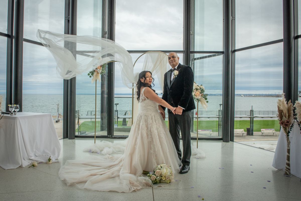 A bride and groom stand in a modern glass-walled space overlooking water, with her veil billowing in the wind.