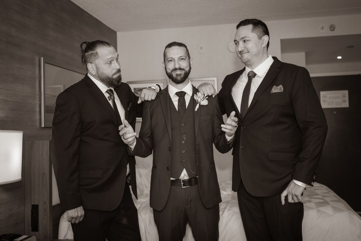 Three men in suits stand close, smiling and gesturing, with the central figure flanked by two groomsmen.