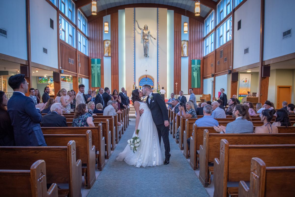 A bride and groom kiss at an altar surrounded by guests in wooden pews, with light streaming through tall windows.