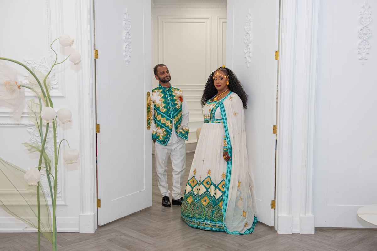 A man and woman in traditional attire stand by an open doorway in a decorated room, bathed in natural light.