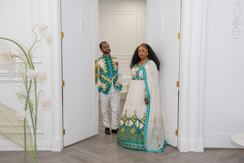 A man and woman in traditional attire stand by an open doorway in a decorated room, bathed in natural light.