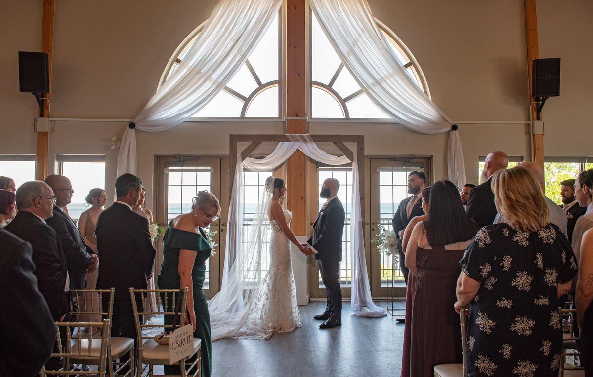 Bride and groom exchange vows under a wooden arch with natural light streaming in through arched windows, guests seated.