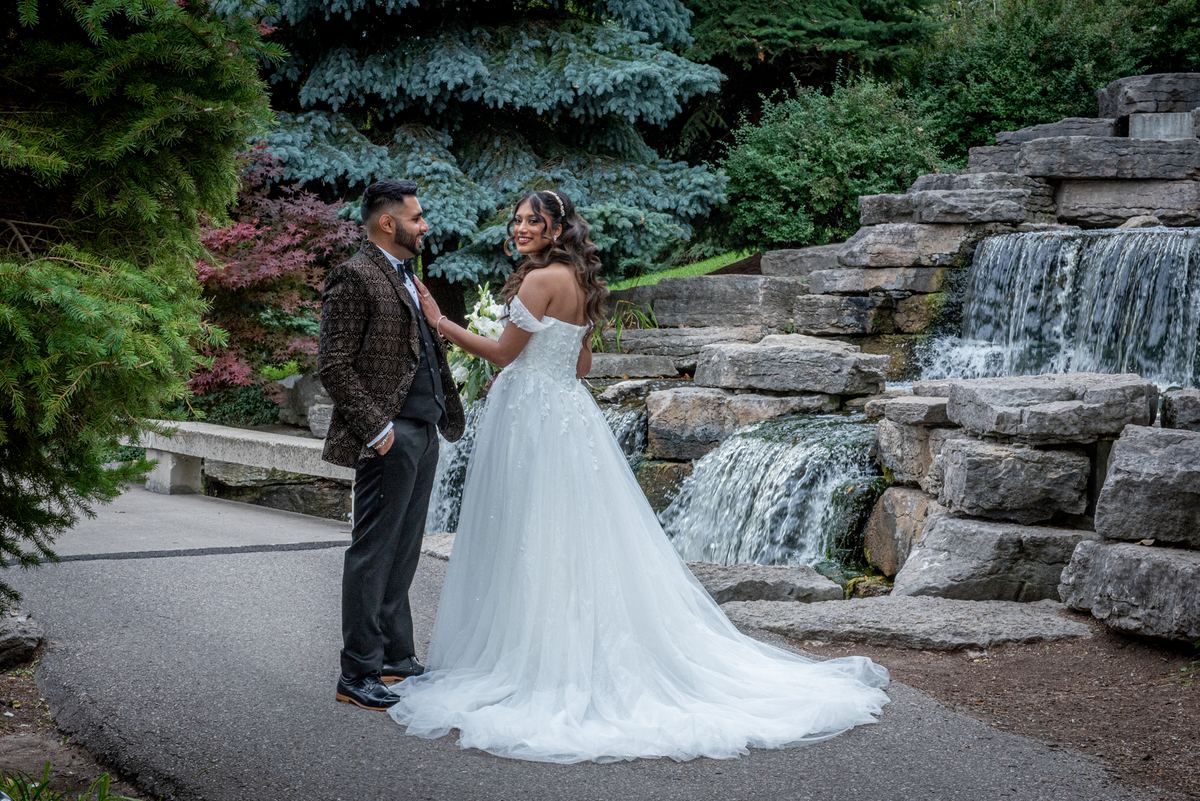 A newlywed couple stands by a stone waterfall, the bride in a white gown touching the groom's chest as he smiles.