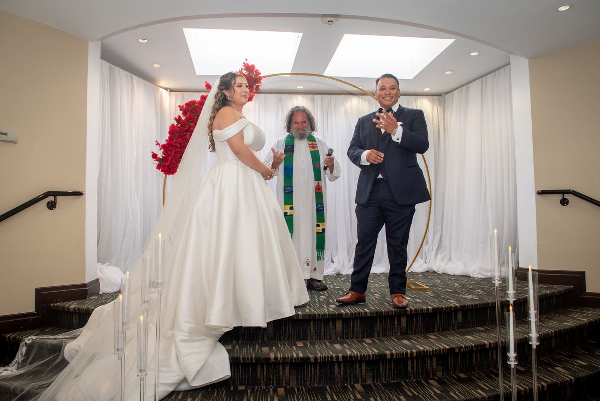 Bride in white gown with red flowers stands beside groom in dark suit on tiered stage during ceremony.