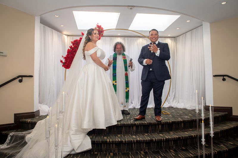 Bride in white gown with red flowers stands beside groom in dark suit on tiered stage during ceremony.