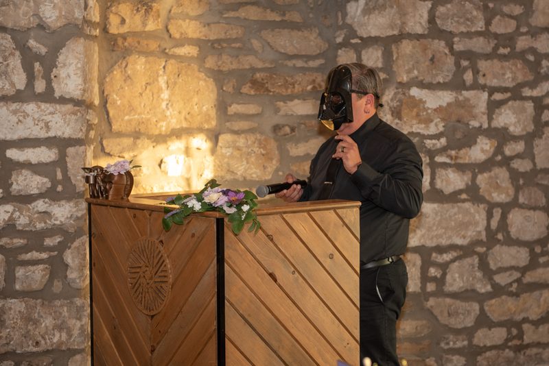 A man in a Darth Vader mask stands at a wooden podium against a warmly lit stone wall, holding a microphone.