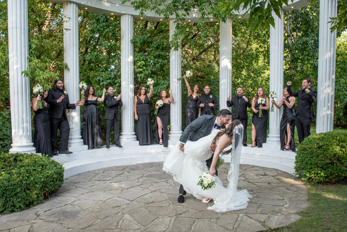 Groom dips bride playfully as wedding party cheers and raises bouquets around white colonnade bathed in daylight.