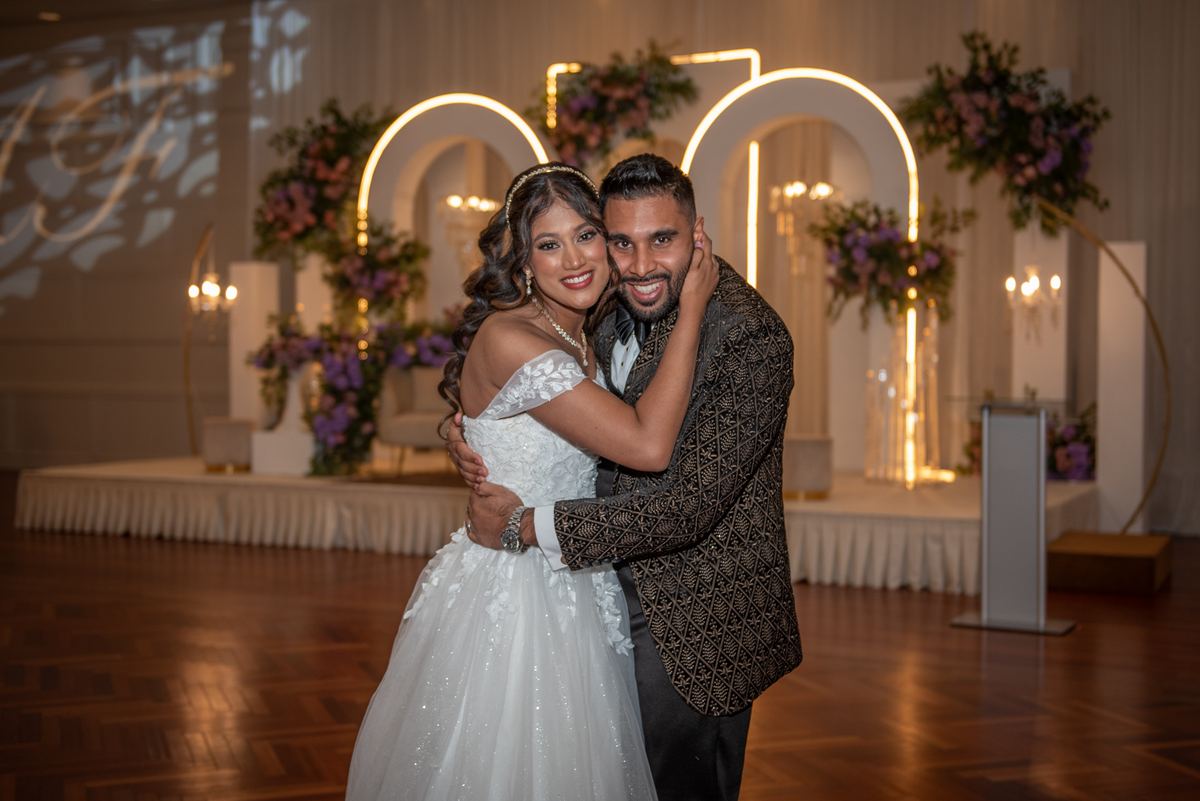 A smiling bride and groom embrace on a dance floor illuminated by glowing arches and floral arrangements.