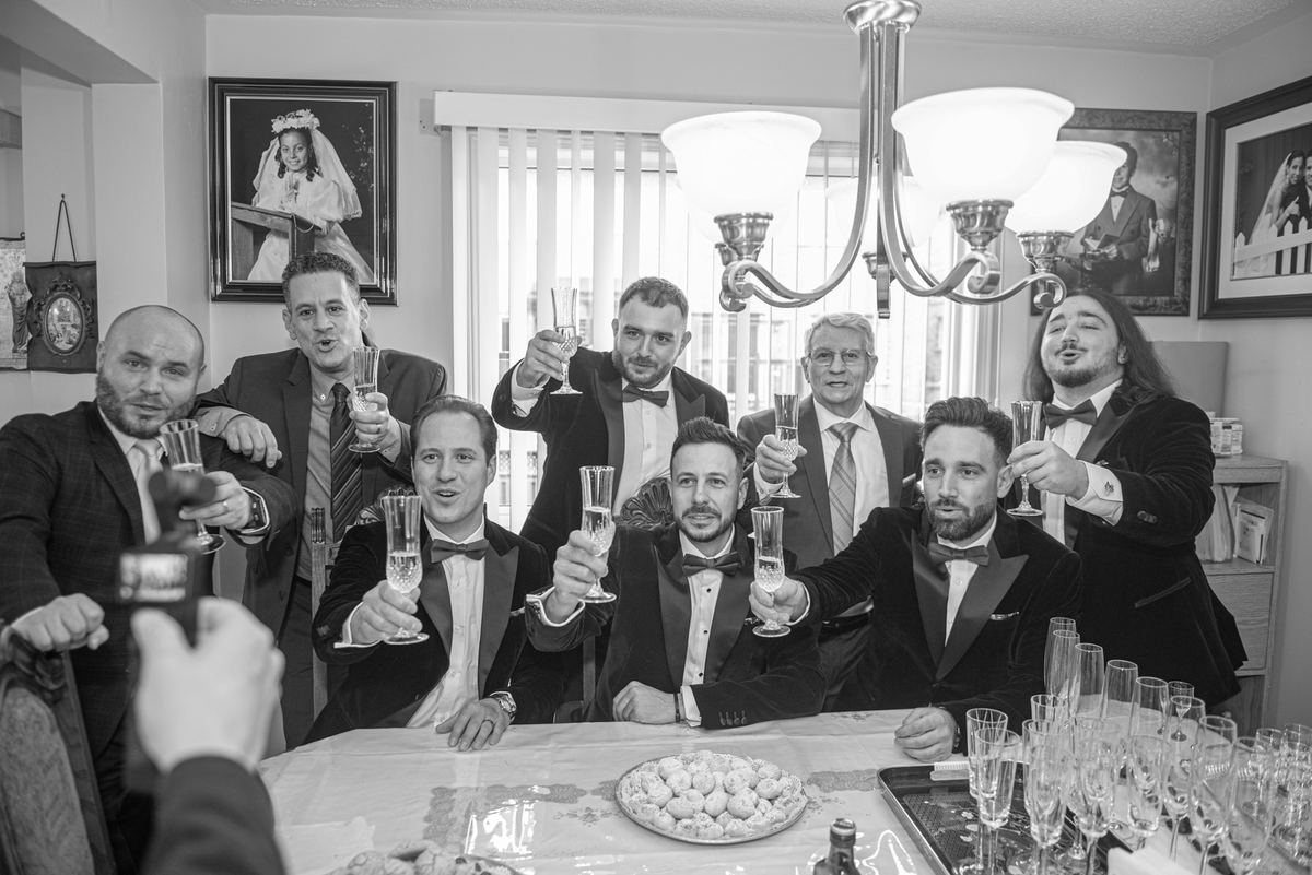 Men in tuxedos and suits gather around a table, raising champagne flutes under a chandelier's glow, with pastries.