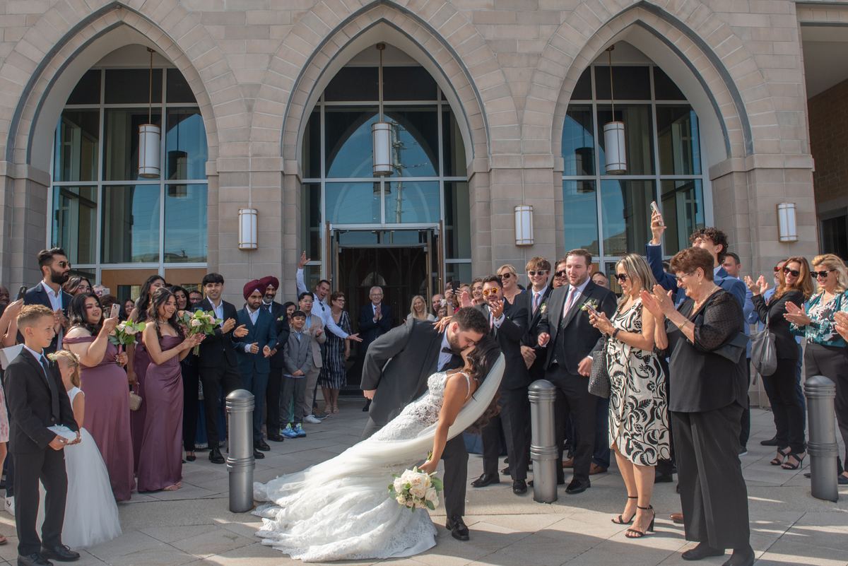A bride and groom kiss joyfully outside a stone building as cheering guests applaud with their phones.