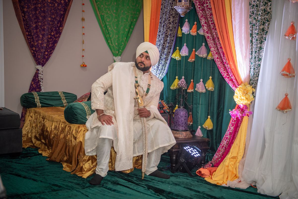 A groom in white ceremonial attire sits thoughtfully on a decorated platform under richly draped fabrics.