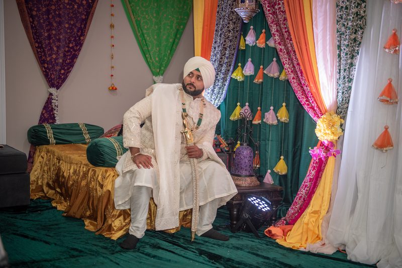 A groom in white ceremonial attire sits thoughtfully on a decorated platform under richly draped fabrics.
