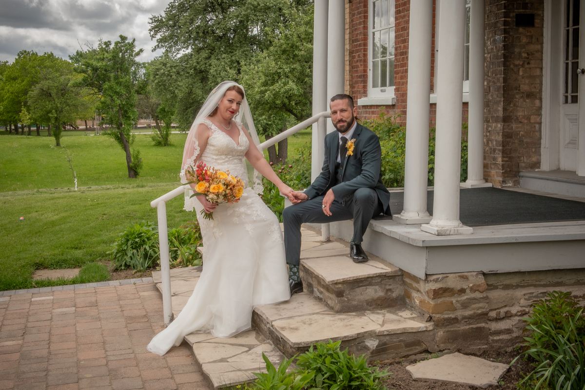 A bride in a white lace gown descends stone steps, holding hands with her groom seated on the porch.