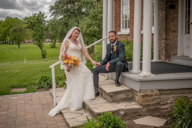 A bride in a white lace gown descends stone steps, holding hands with her groom seated on the porch.