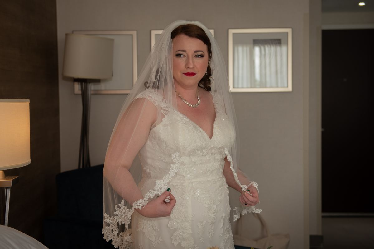 Bride adjusting her veil in a softly lit room, holding the train of her gown, with warm light highlighting lace details.