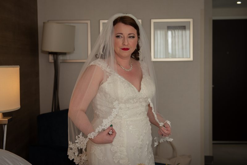 Bride adjusting her veil in a softly lit room, holding the train of her gown, with warm light highlighting lace details.