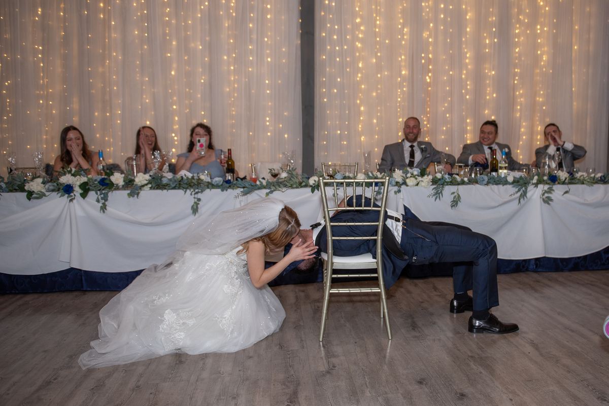 The bride kneels beside her slumbering groom at the reception, where guests laugh and watch, surrounded by fairy lights.