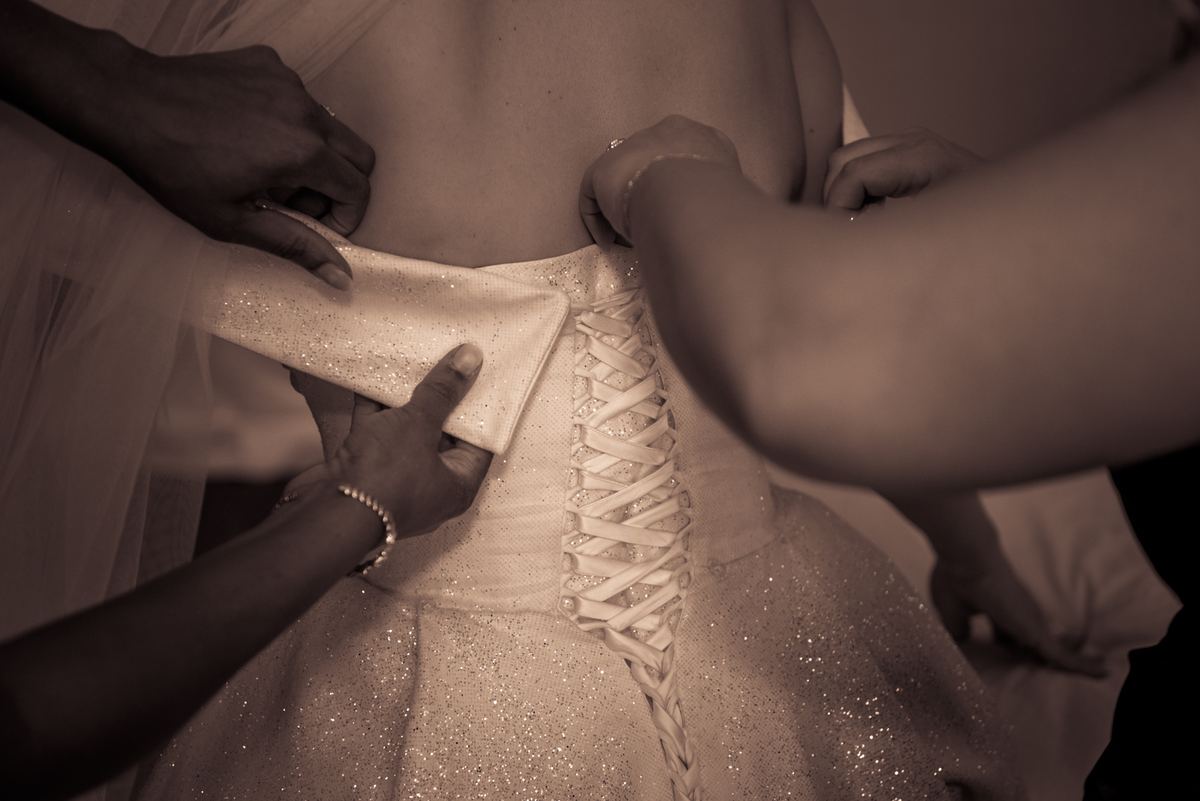 Hands delicately adjust the back of a shimmering wedding gown in warm sepia lighting, emphasizing the fabric's texture.
