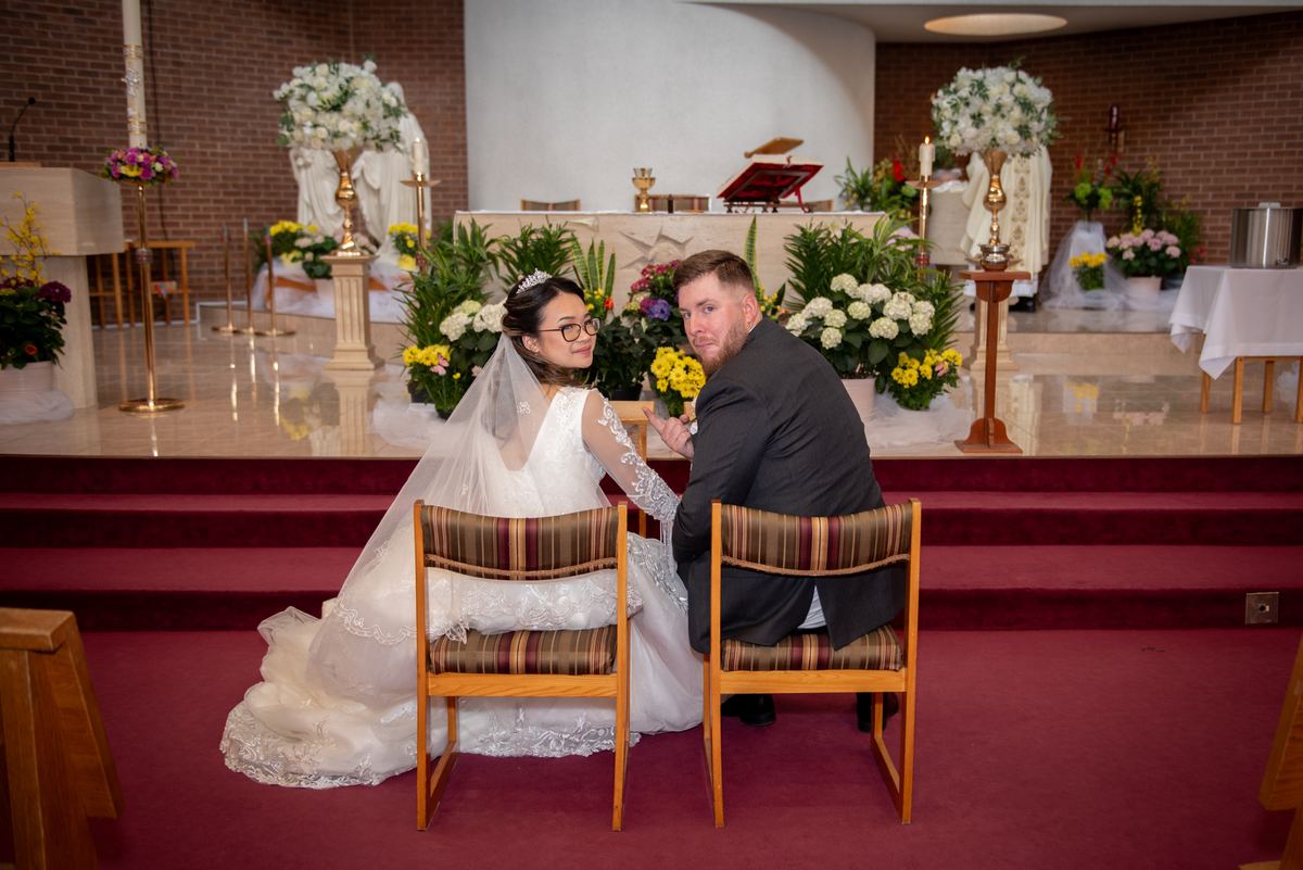 A bride and groom sit together on a red carpet, looking affectionately at each other before their ceremony.