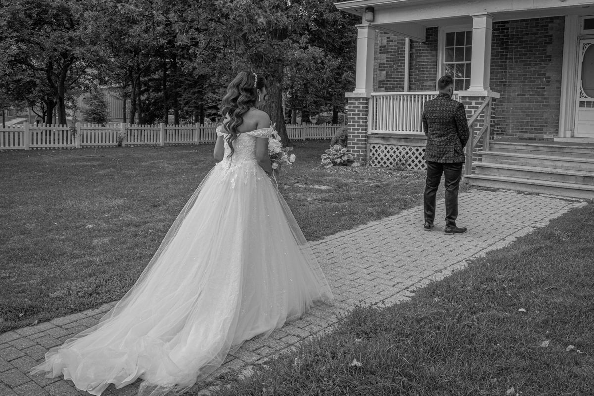 Bride in white dress and groom stand facing each other on steps, awaiting ceremony, in soft black-and-white light.