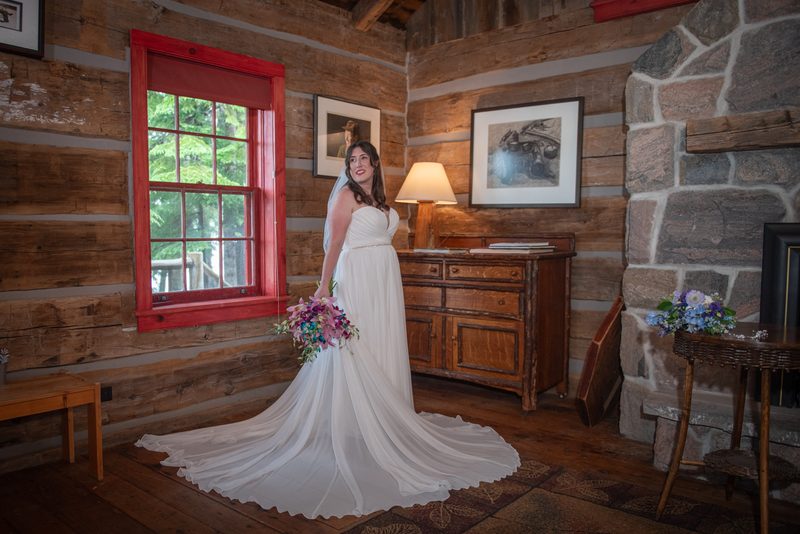 A bride in a flowing white gown stands smiling beside a red-framed window in a rustic log cabin, holding a colorful bouquet.